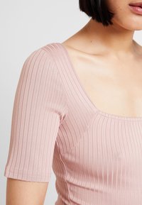 Top côtelé rose clair avec manches courtes et large encolure carrée, confectionné dans un tissu extensible qui met en valeur la silhouette.