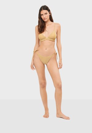 Vrouw staat op blote voeten en draagt een gestructureerde gele bikini met gebonden zijbandjes tegen een effen lichte achtergrond.