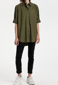 Camicia verde oliva con maniche corte arrotolate e taschino sul petto, abbinata a pantaloni neri slim-fit e sneakers bianche.