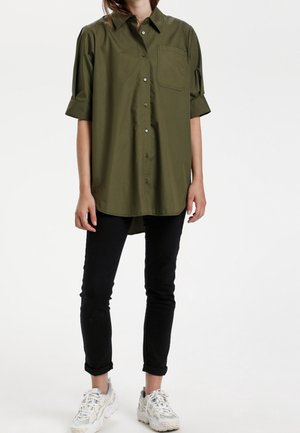 Chemise boutonnée vert olive à manches courtes retroussées et poche poitrine, associée à un pantalon noir ajusté et des baskets blanches.