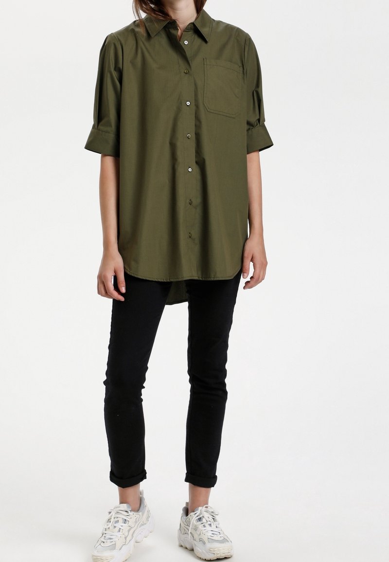 Camicia verde oliva con maniche corte arrotolate e taschino sul petto, abbinata a pantaloni neri slim-fit e sneakers bianche.