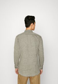 Camisa de manga larga gris con tela texturizada, puños abotonados, canesú en la parte posterior y dobladillo curvado. Usada por una persona, vista desde la espalda.