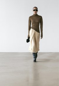 Bruine top met lange mouwen, crèmekleurige wijdvallende broek met geplooide stof en zwarte laarzen tot over de knie. Accessoires zijn een kleine zwarte tas en een zonnebril.