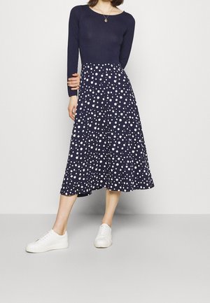 Kvinde iført navyblå langærmet top og navyblå midinederdel med hvide polkaprikker, kombineret med hvide sneakers, står ved en ensfarvet væg.
