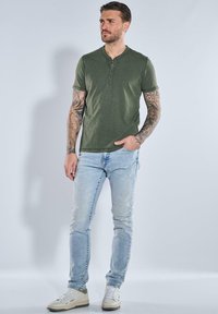 Olive green, short-sleeve henley t-shirt with a button placket, paired with light blue, slim-fit jeans and white sneakers. Tattoos on forearms visible.
