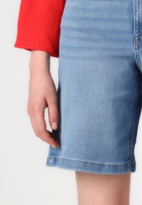 Jasnoniebieskie szorty dżinsowe o prostym kroju i postrzępionych końcach, noszone z czerwoną, fakturowaną rękawicą widoczną na lewej dłoni.