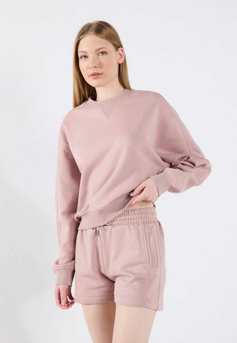Sudadera rosa claro con cuello redondo y corte relajado, combinada con pantalones cortos a juego que tienen una cinturilla elástica y bolsillos laterales.