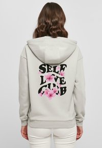 Felpa grigia chiara con tasca frontale, con la scritta "SELF LOVE CLUB" in grassetto nero, accentuata da grafiche di fiori rosa.