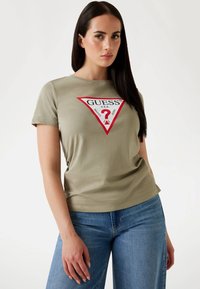 T-shirt khaki a maniche corte con una grafica a triangolo rosso e il testo "GUESS" sopra un punto interrogativo e dettagli sui jeans. Vestibilità casual.