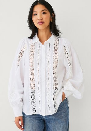 Kobieta ubrana w białą bluzkę z długim rękawem z koronkowymi panelami i bufiastymi rękawami, w połączeniu z niebieskimi dżinsami, stojąca na jednolitym tle.