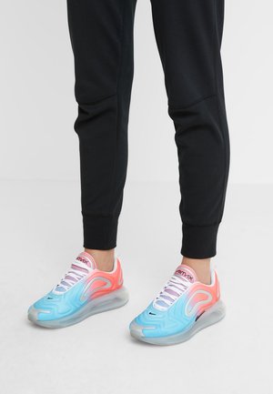 Personne portant un pantalon noir et des baskets Nike Air Max colorées bleues et roses debout sur un sol blanc.