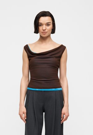 Νεαρή γυναίκα με κοντά σκοτεινά μαλλιά φοράει καφέ τοπ με λαιμόκοψη πάνω από μπλε πουκάμισο και σκούρο γκρι πλισέ παντελόνι, πόζαρε μπροστά σε λευκό φόντο.