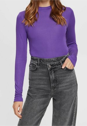 Femme portant un haut violet à manches longues rentré dans un jean noir délavé taille haute, une main dans la poche et des cheveux lâches brun-roux.