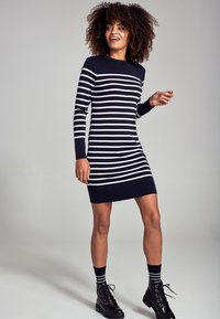 Robe rayée à manches longues, couleurs marine et blanche, texture côtelée, longueur genou. Associée à des chaussettes jusqu'aux genoux et des bottes noires à lacets.
