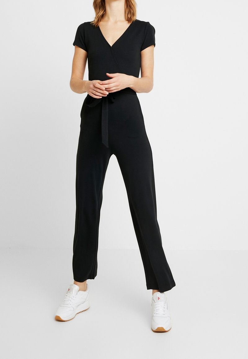 Combinaison noire à manches courtes avec décolleté en V, taille lien, coupe décontractée et jambes droites, fabriquée en tissu lisse. Baskets blanches avec une semelle texturée.