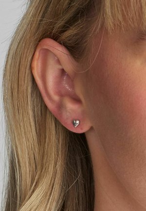 Srebrna naušnica u obliku srca pričvršćena na uho, s poliranom površinom i malom veličinom, s teksturiranim rubom za dodatne detalje.