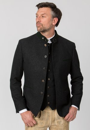 Man wearing dark traditional jacket over black vest and white shirt, paired with beige embroidered trousers, looking to side against plain background.