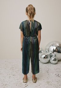 Tuta brillante e multicolore con texture plissettata, maniche corte e schiena a chiave. Abbinata a calzature dorate, sullo sfondo di sfere da discoteca.