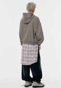Felpa grigia oversize con cappuccio, indossata sopra una tunica a quadri rosa, abbinata a pantaloni neri larghi e calzature beige con texture.