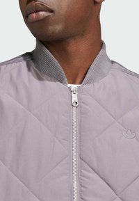 Vadderad ljuslila bomberjacka med ribbad krage, silverfärgad dragkedja och broderad logotyp på bröstet. Slät tygtextur.