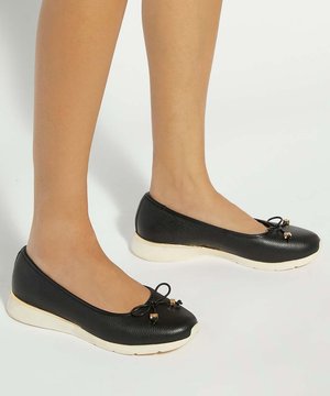 Beine, die schwarze Leder-Ballerinas mit kleinen Schleifen und weißen Sohlen auf hellem Hintergrund tragen.