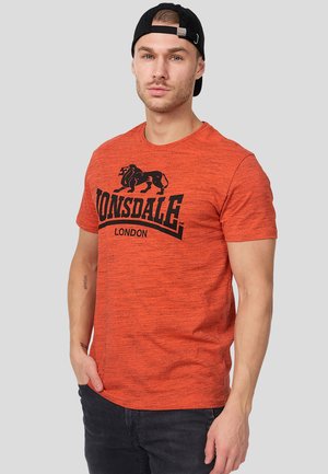 Mand iført en sort kasket bagud og en orange Lonsdale London t-shirt, stående mod en ensartet lysegrå baggrund.