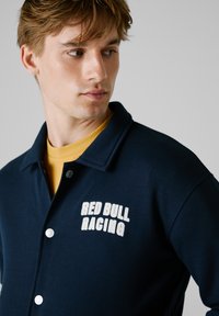 Marineblå knap-jerseyjakke lavet af blødt stof, med et broderet "RED BULL RACING" logo i hvid på venstre side.