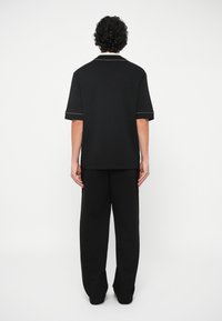 Άνδρας που στέκεται με την πλάτη γυρισμένη, φορώντας μαύρο κοντομάνικο πουκάμισο και φαρδιά μαύρα παντελόνια, μπροστά σε απλό λευκό φόντο.