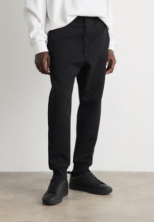 Mann trägt schwarze Joggerhose mit Kordelzug und schwarze Sneakers, kombiniert mit einem weißen Langarmshirt, steht auf Betonboden.