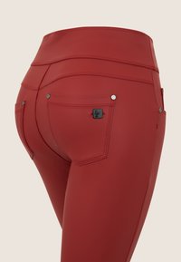 Leggings rossi a vita alta con una texture liscia e setosa. Presentano due tasche posteriori con finiture in metallo argentato e un piccolo etichetta con marchio.