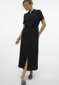 Robe noire à manches courtes et à boutons, avec une taille nouée et une fente sur le côté, fabriquée dans un tissu lisse. Les accessoires comprennent un grand bracelet et des sandales bleues.