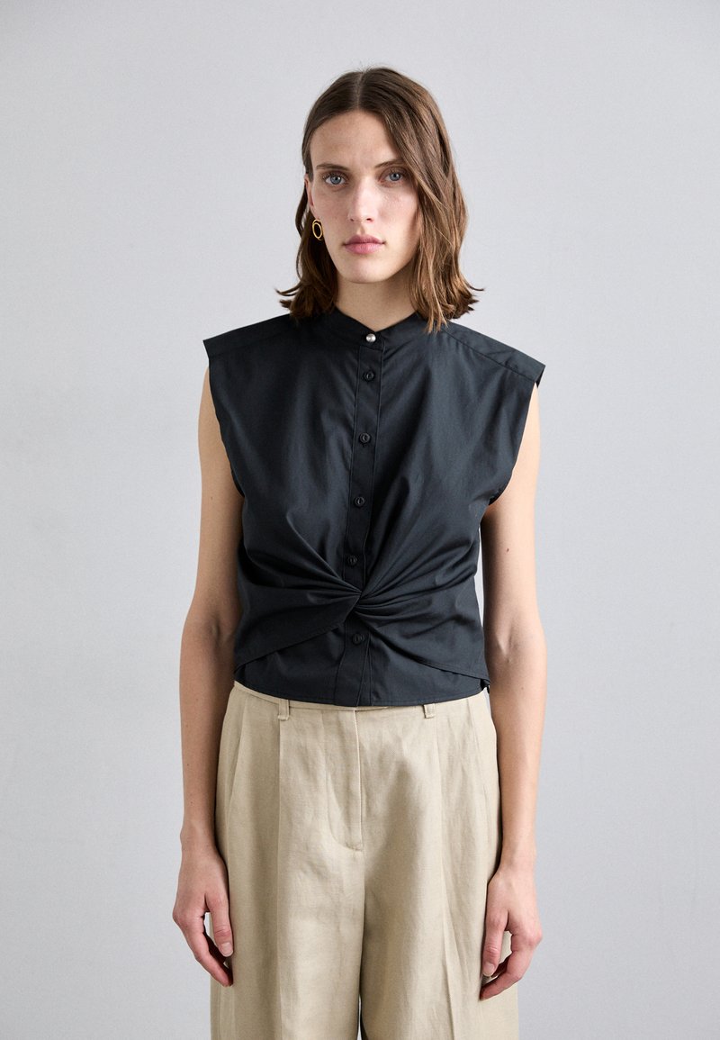 Cămașă fără mâneci, neagră, cu un design legat la talie, asortată cu pantaloni evazați de un bej deschis. Material neted, stil minimalist.