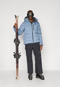 Svijetloplavi puffer jakna, crne ski hlače i ski čizme. Drži drvene skije. Dodaci uključuju naočale i jaknu s patentnim zatvaračem.