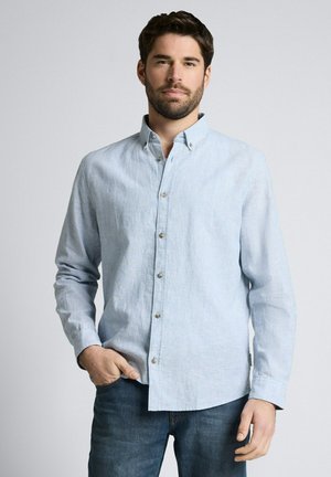 Man with dark hair and beard wearing light blue button-up shirt and dark jeans, standing with one hand in pocket against plain background.