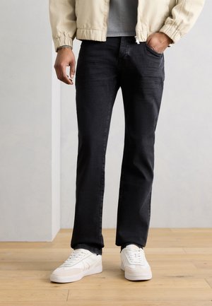 Personne portant un jean noir, des baskets blanches, une chemise grise et une veste beige, debout sur un sol en bois devant un mur clair uni.