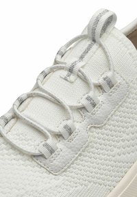 Sneaker bianca con tomaia in tessuto testurizzato, lacci elastici e dettagli grigi sui fori per i lacci. La suola è spessa e di tonalità neutra.