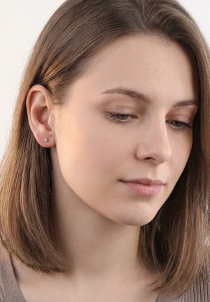 Jeune femme aux cheveux lisses châtain clair, portant de petites boucles d'oreilles rondes, regardant vers le bas avec une expression neutre.