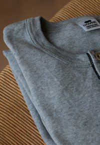Grå bomulds sweater foldet, med en rund hals, et mærke med "DOVRE" og en knapdetalje nær halsen, på en vævet overflade.