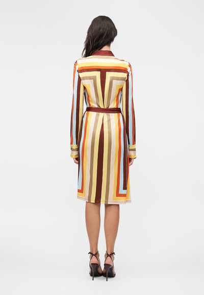 Γυναίκα όρθια γυρισμένη με την πλάτη, φοράει φόρεμα μέχρι το γόνατο με κάθετες πολύχρωμες ρίγες σε καφέ, πορτοκαλί, κίτρινο, μπλε και μπεζ, συνδυασμένο με μαύρα τακούνια.