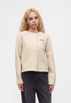 Jeune femme aux longs cheveux blonds bouclés portant une veste polaire crème Columbia et un pantalon foncé, debout devant un fond blanc uni.