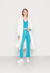 Casaco longo branco sobre um top cropped turquesa com decote drapeado e calças largas combinando, acompanhado por ténis brancos e uma mala pequena.