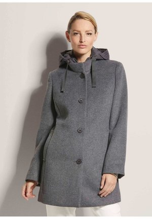 Femme portant un manteau en laine gris mi-long avec des boutons noirs et une capuche avec des cordons noirs, debout devant un fond neutre.