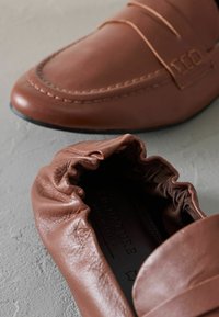 Mocassini in pelle marrone con dettagli cuciti, caratterizzati da una texture arricciata e da una silhouette slanciata. Suole in gomma nera visibili.