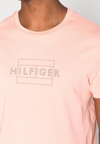 Camiseta de algodón color rosa claro con mangas cortas, cuello redondo y un logo "HILFIGER" impreso en negro delineado en la parte delantera.