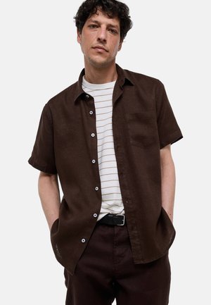 Jeune homme portant une chemise marron foncé à manches courtes boutonnée par-dessus un t-shirt blanc rayé, les mains dans les poches, debout devant un fond uni.