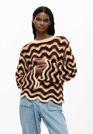 Kvinde iført en flerfarvet, bølget strikket sweater med bølgekant og lyseblå højtragtede jeans, stående mod en hvid baggrund.