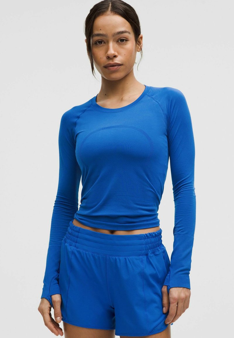 Sinine pika varrukaga sportlik topp, millele on kombineeritud sobivad lühikesed püksid. Mõlemad on valmistatud kergest, venivast kangast, millel on sile tekstuur ja sobiv disain.