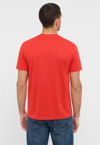 Czerwona koszulka z krótkim rękawem i okrągłym dekoltem, o gładkiej fakturze i minimalnych detalach, zestawiona z niebieskimi dżinsami posiadającymi tylne kieszenie.