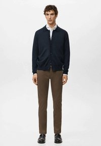 Granatowy dzianinowy kardigan na guziki, noszony na białej koszuli z kołnierzykiem, zestawiony z brązowymi wąskimi spodniami i czarnymi wsuwanymi butami.