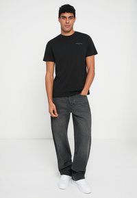 Μαύρο βαμβακερό μπλουζάκι με στρογγυλή λαιμόκοψη και μικρό λογότυπο, συνδυασμένο με φαρδιά γκρίζα τζιν παντελόνια και λευκά αθλητικά παπούτσια. Απλός, casual σχεδιασμός.